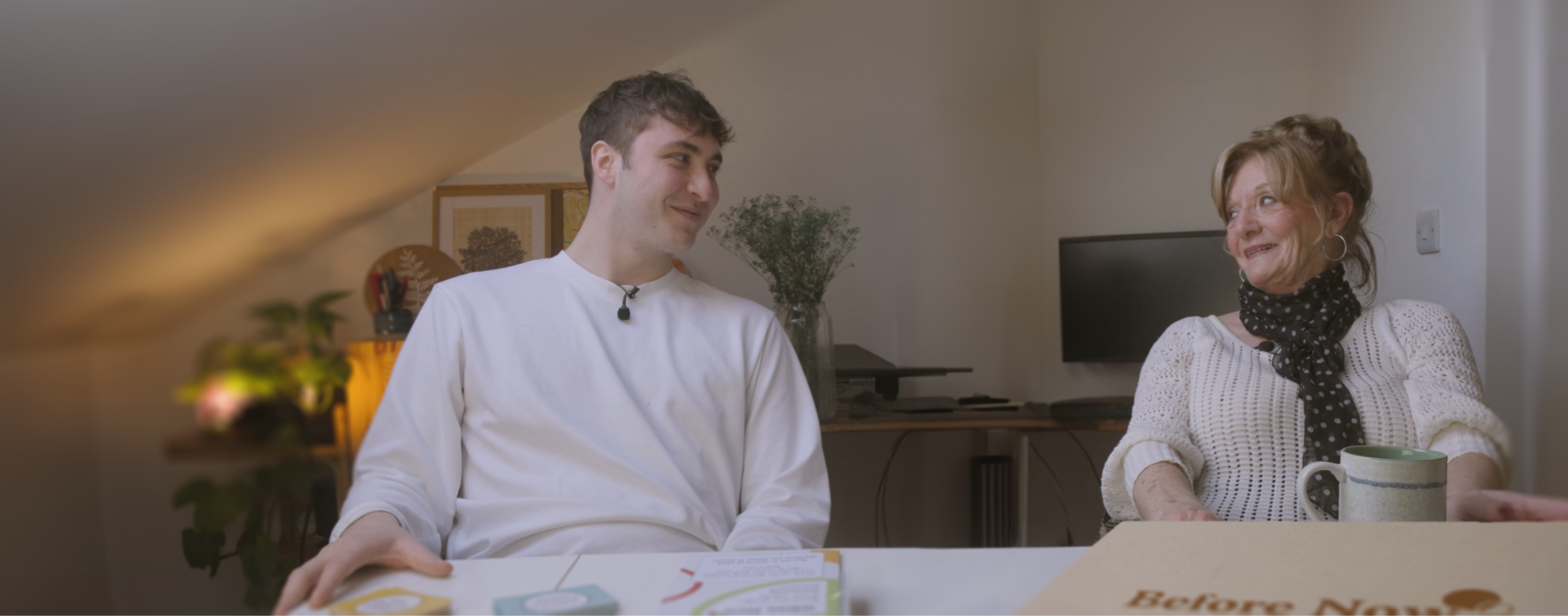 A grandson and grandmother smiling at each other while using Before Now, the life history collection kit that brings people together through conversation. Help loved ones remember their lives, talk to your older relatives and friends using conversation cards that prompt meaningful discussions, and turn your recordings into a guided autobiograhy.